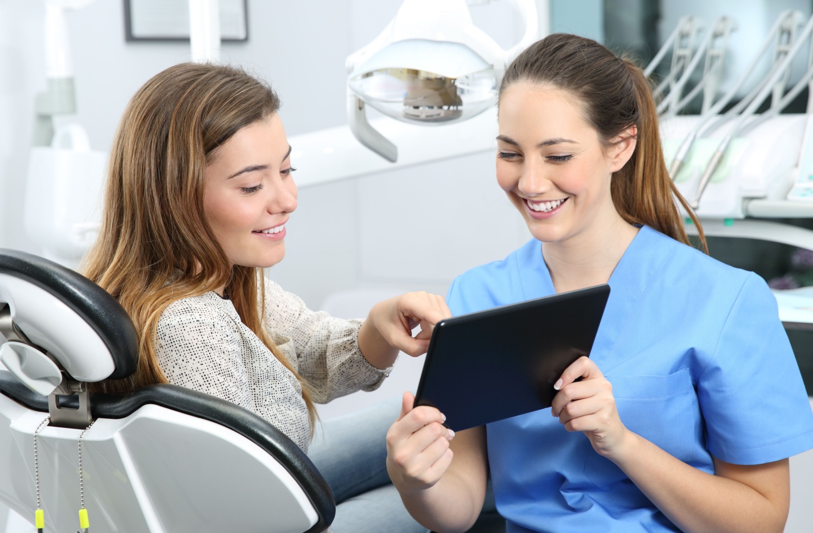 A dental hygienist and a patient sitting in a dental chair smiling while looking at information on a tablet.